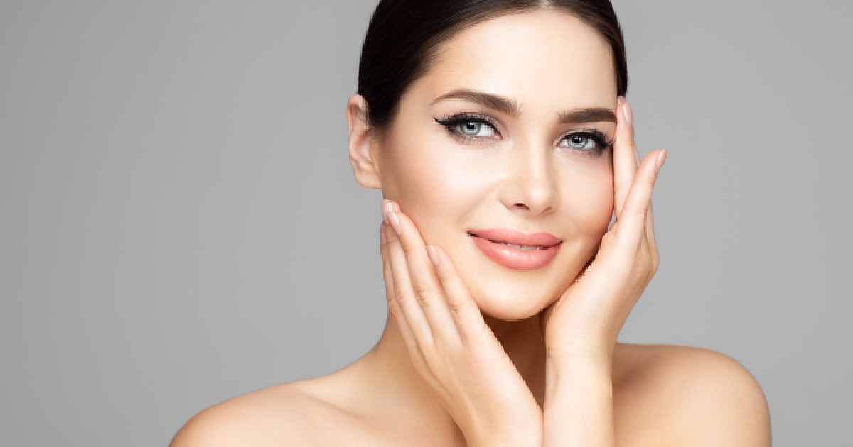 Beautiful woman with flawless glowing skin after a Signature Facial in Washington, DC, with subtle makeup and a soft smile, gently touching her face with both hands against a neutral gray background.