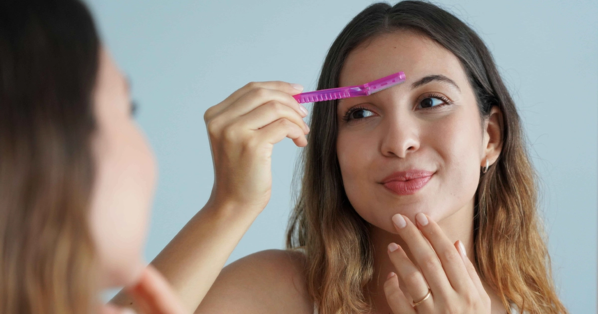 Woman using a pink facial razor for Dermaplaning in Washington, DC to groom her eyebrows while looking in a mirror.